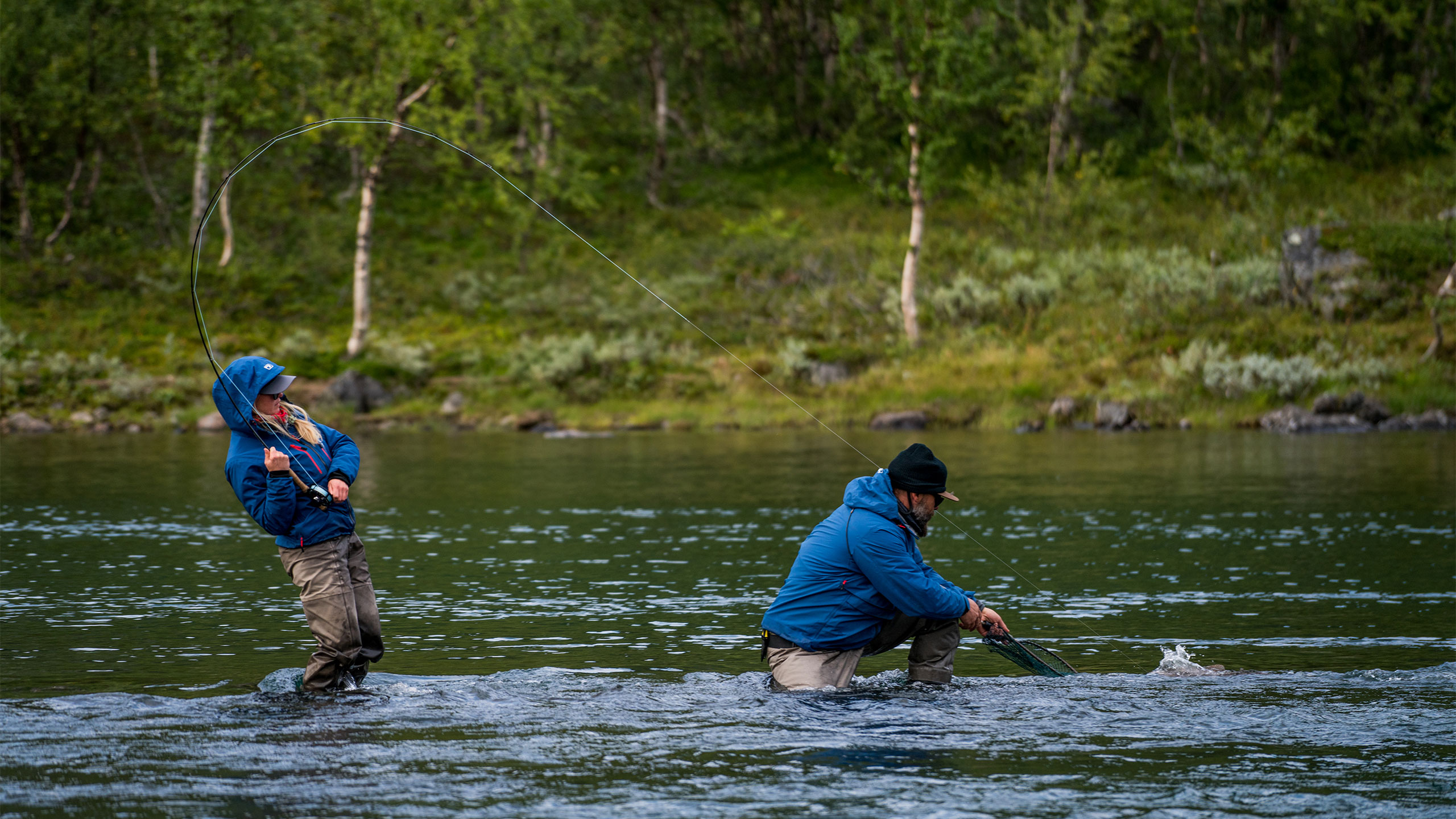 To fiskere vader i elva, den ene håver inn fisk